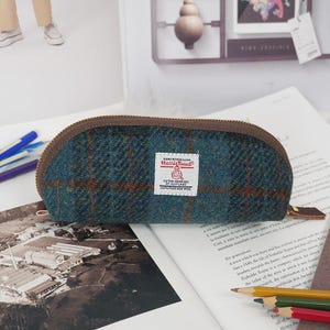 May include: A semi-circular pencil case made of blue and green Harris Tweed wool fabric with a brown plaid pattern. The case features a brown zipper closure and a white rectangular label with red text reading Harris Tweed Outer Hebrides.