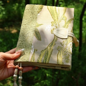 Puede incluir: Un diario decorativo con una cubierta crema con un estampado floral verde y un borde de encaje verde claro. El diario tiene un cierre de cuero crema y una borla con cuentas de madera.