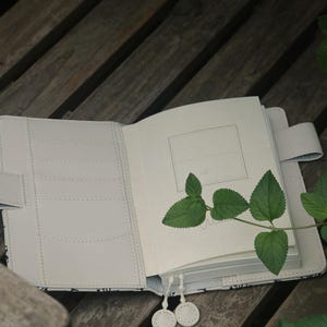 Op de afbeelding: Een wit lederen gebonden dagboek met een zwart-wit patroon op de omslag. Het dagboek ligt open op een blanco pagina met een groene plantentak erop.