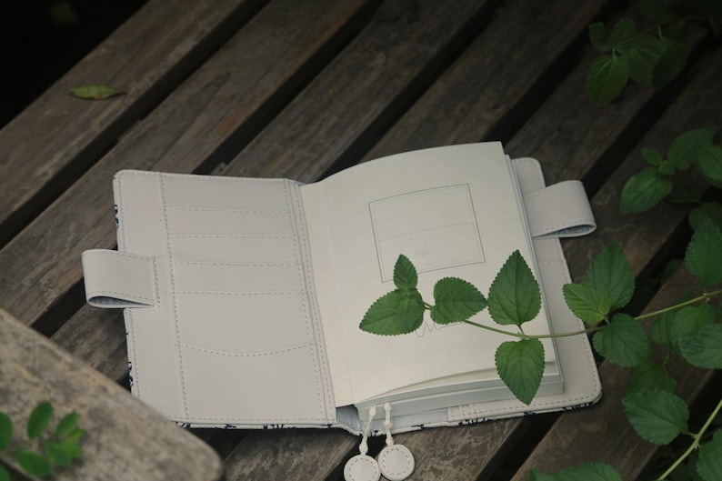 Op de afbeelding: Een wit lederen gebonden dagboek met een zwart-wit patroon op de omslag. Het dagboek ligt open op een blanco pagina met een groene plantentak erop.