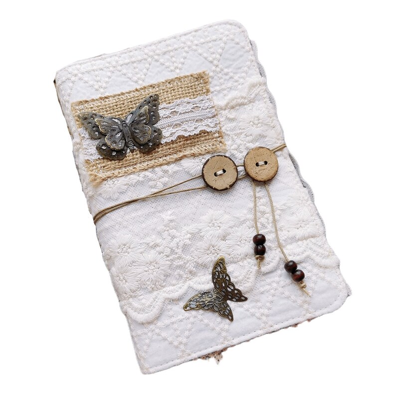 May include: A white fabric journal cover with a burlap and lace overlay. The cover features two metal butterfly embellishments, two wooden buttons, and brown beads on strings.