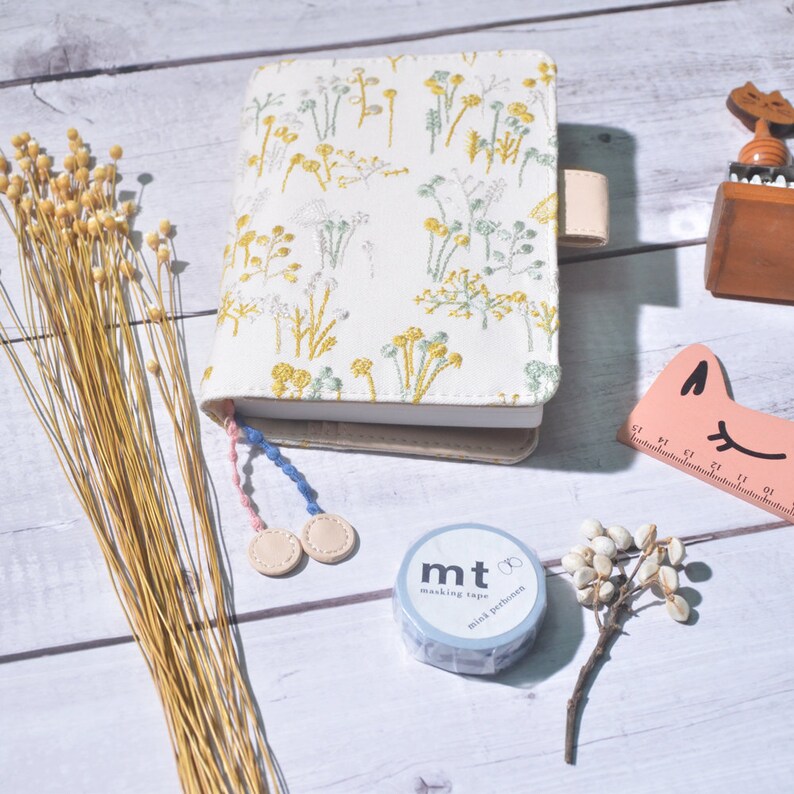 May include: A white notebook cover with a floral embroidery design in yellow and green. The cover has a beige strap and a pink and blue tassel. A roll of blue washi tape with the text "mt masking tape mini pattern" is visible in the foreground.