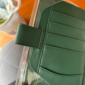 May include: A dark green leather planner with a clear plastic cover and multiple pockets. The planner has a flap closure with a black strap.