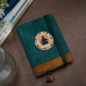 Puede incluir: Un diario de pana verde azulado con un parche bordado decorativo con un diseño de casa. El diario tiene una banda elástica y un borde marrón, y un pequeño amuleto en forma de calabaza atado a una cinta.
