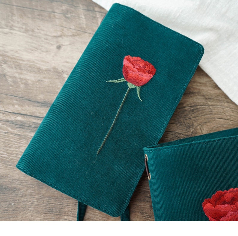 May include: A green corduroy notebook with a red embroidered rose. The notebook has a simple design and is perfect for journaling or taking notes.