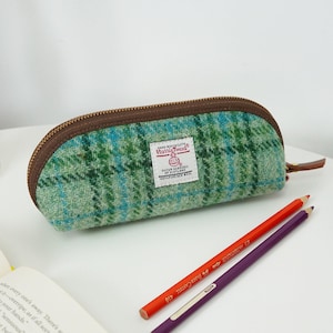 May include: A green and blue plaid pencil case made of Harris Tweed wool fabric with a brown zipper closure. A white rectangular label on the front features the text Harris Tweed Hand Woven Cloth Outer Hebrides of Scotland 100% Pure New Wool.