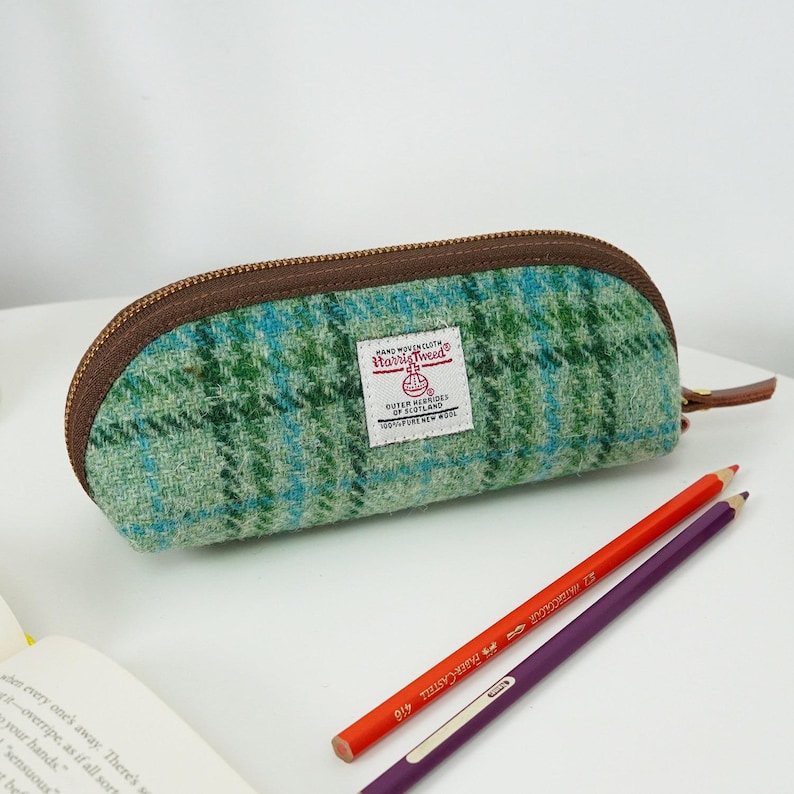 May include: A green and blue plaid pencil case made of Harris Tweed wool fabric with a brown zipper closure. A white rectangular label on the front features the text Harris Tweed Hand Woven Cloth Outer Hebrides of Scotland 100% Pure New Wool.