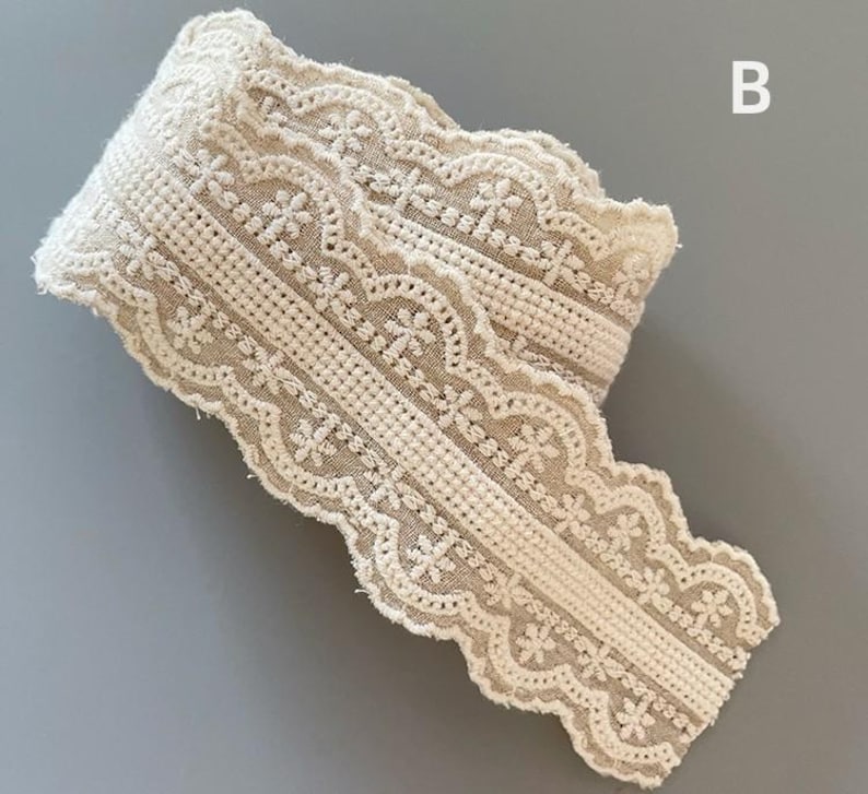May include: A close-up of a beige crocheted lace trim with a scalloped edge. The lace has a delicate floral pattern.