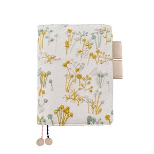 May include: A white fabric notebook cover with a floral embroidery design in yellow and green. The cover has two beige leather straps and a pink and blue tassel.