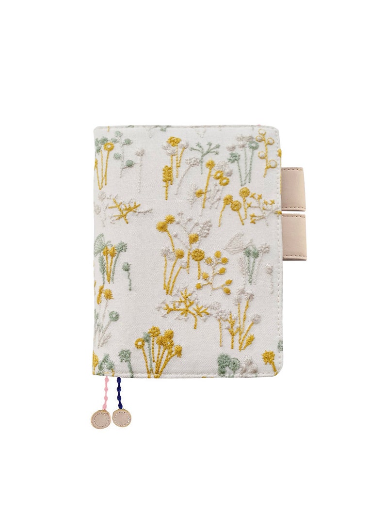 May include: A white fabric notebook cover with a floral embroidery design in yellow and green. The cover has two beige leather straps and a pink and blue tassel.