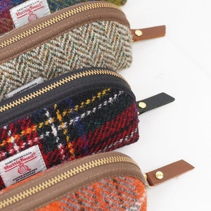 May include: Three zippered pouches made of Harris Tweed wool fabric. The top pouch features a green and brown herringbone pattern with a brown zipper and leather pull. The middle pouch has a red, black, and yellow plaid pattern with a black zipper and leather pull. The bottom pouch is orange with a brown zipper and leather pull.