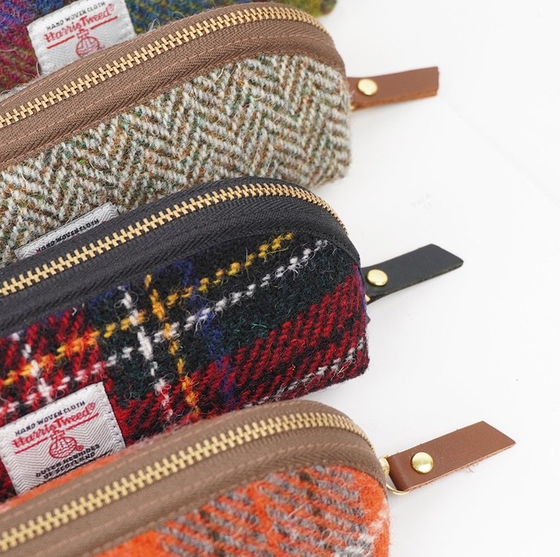 May include: Three zippered pouches made of Harris Tweed wool fabric. The top pouch features a green and brown herringbone pattern with a brown zipper and leather pull. The middle pouch has a red, black, and yellow plaid pattern with a black zipper and leather pull. The bottom pouch is orange with a brown zipper and leather pull.