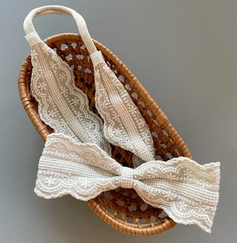 May include: A white lace headband with a bow. The headband is in a woven basket.