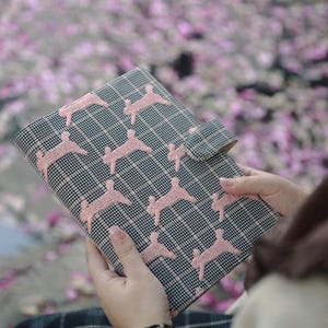 May include: A black and white checked binder with pink embroidered designs. The binder has a flap closure and is being held in someone's hands.