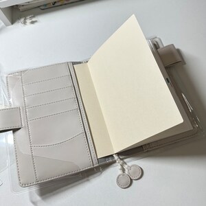 May include: A gray leather bound journal with a clear plastic cover and a white ribbon bookmark. The journal is open to a blank page.