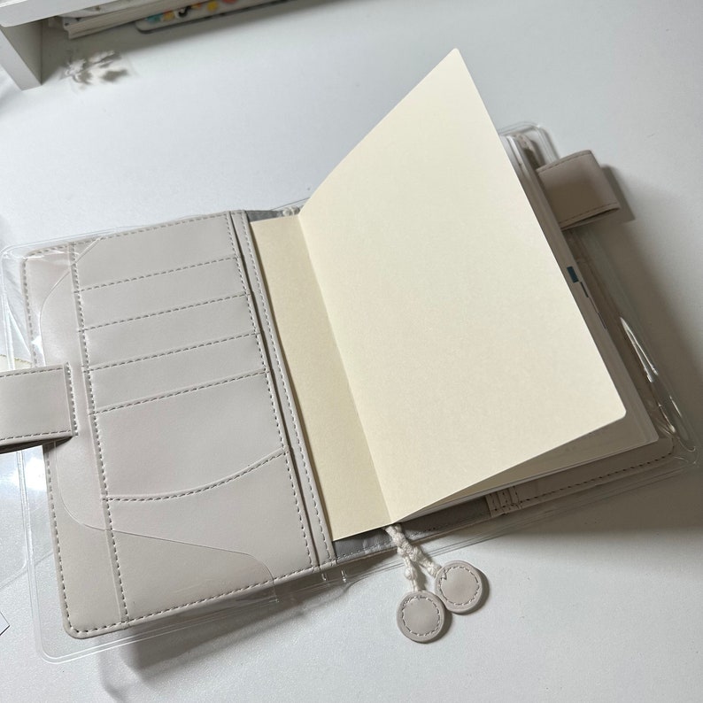 May include: A gray leather bound journal with a clear plastic cover and a white ribbon bookmark. The journal is open to a blank page.