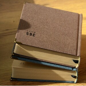 May include: Three hardcover journals with brown, blue, and brown covers. The covers are textured and have gold corner accents. The journals are stacked on top of each other. The top journal has the text "365 DAYS" printed on the cover. The middle journal has the text "365 DAYS" printed on the cover. The bottom journal has the text "365 DAYS" printed on the cover.