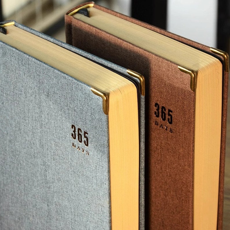 May include: Two hardcover journals with gold accents and the text "365 Days" on the front. The journal on the left is gray and the journal on the right is brown.