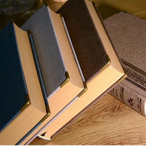 以下が含まれることがあります： 青、グレー、茶色など、さまざまな色の表紙が付いたハードカバーの本のコレクション。各本には金色のコーナープロテクターと明るい色のページが付いています。本は木製の表面に配置されています。