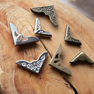 Könnte beinhalten: Eine Auswahl an dekorativen Metallecken in Silber- und Antik-Messing-Ausführungen. Die Ecken weisen verzierte, gerollte Designs auf und sind auf einer Holzoberfläche angeordnet. Ideal für Scrapbooking und andere Bastelarbeiten. Die Ecken sind aus Metall.
