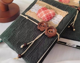 May include: A dark green fabric-covered journal with a burlap and lace accent. The journal has a red and white plaid fabric apple, two wooden buttons, and a brown twine closure.