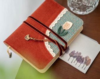Puede incluir: Un diario hecho a mano con una cubierta de pana color óxido y una sección estampada en verde azulado y blanco. El diario está asegurado con una cinta de terciopelo rojo oscuro y presenta una aplicación decorativa de tulipán. Una pequeña tarjeta con ilustraciones de árboles descansa a su lado.