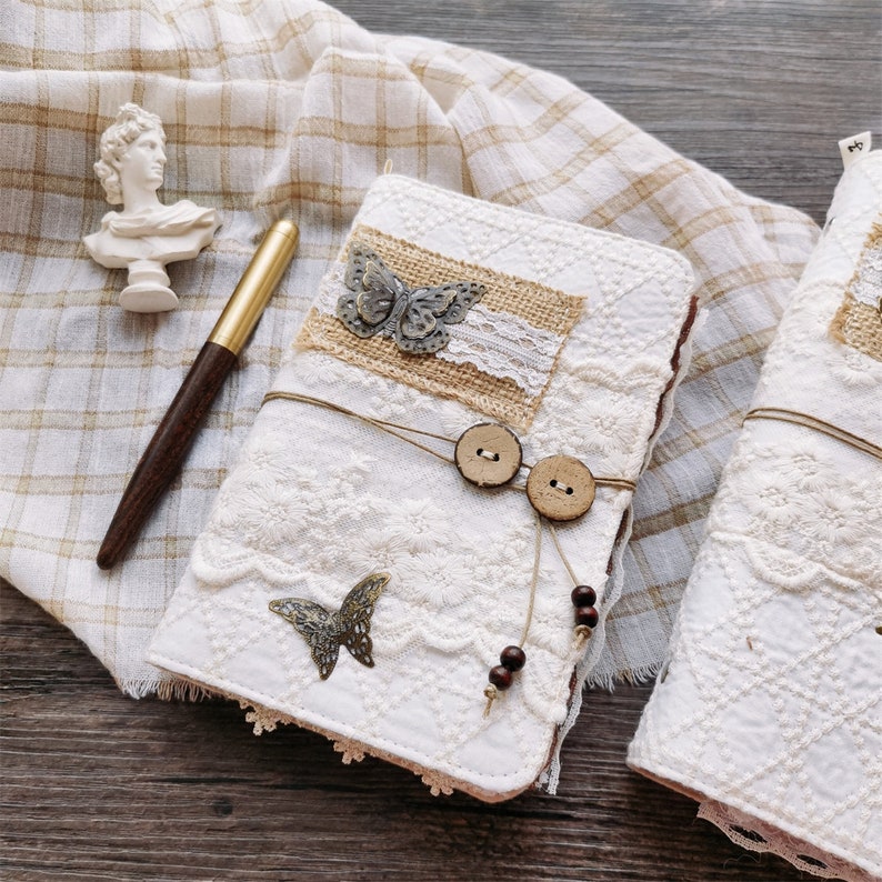 May include: Two white fabric journals with lace and burlap accents. The journals have butterfly embellishments, wooden buttons, and brown beads. One journal is open to reveal a blank page.