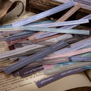 May include: A collection of colorful paper strips with various inspirational quotes and phrases. The strips are printed with different fonts and colors, including black, white, gray, pink, blue, and green. Some of the phrases include "work in just", "be kind and generous", "how empty science", "late spring wealth", and "no company".