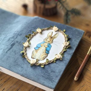 Könnte beinhalten: Ein mit blauem Samt bezogenes Buch mit einer ovalen Illustration eines Kaninchens in einer blauen Jacke. Die Illustration ist von einer geflochtenen Kordel umrahmt, die mit kleinen cremefarbenen Stoffrosen und grünen Blättern verziert ist.