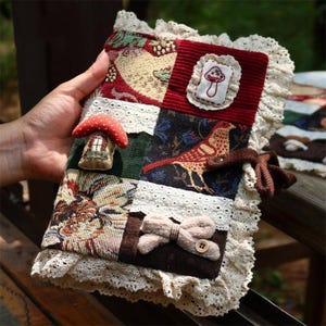 May include: A fabric-covered journal featuring a patchwork design of floral, bird, and mushroom motifs. The cover includes a 3D mushroom embellishment, a small embroidered mushroom patch, a fabric bow with a button, and cream-colored lace trim.