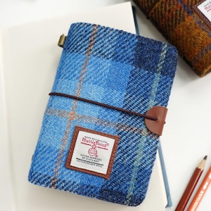 May include: A blue plaid Harris Tweed notebook cover featuring a brown elastic closure band. A white rectangular label on the front displays the text Harris Tweed, Hand Woven Cloth, Outer Hebrides of Scotland, and 100% Pure New Wool.