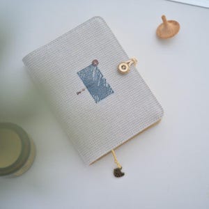Pode incluir: Um diário cinza claro com um design azul e branco e o texto "Day-by-". O diário tem um fecho de botão de madeira e um pequeno encanto. Um pião de madeira também é visível.