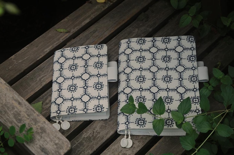 Op de afbeelding: Twee witte, met stof beklede notitieboekjes met blauwe geometrische patronen. De notitieboekjes hebben witte elastische sluitingen en witte kwastaccenten.