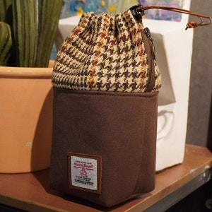 May include: A brown and beige tweed drawstring bag with a zipper closure and a brown canvas base. The bag features a brown leather patch with the text "Harris Tweed" and a small logo.