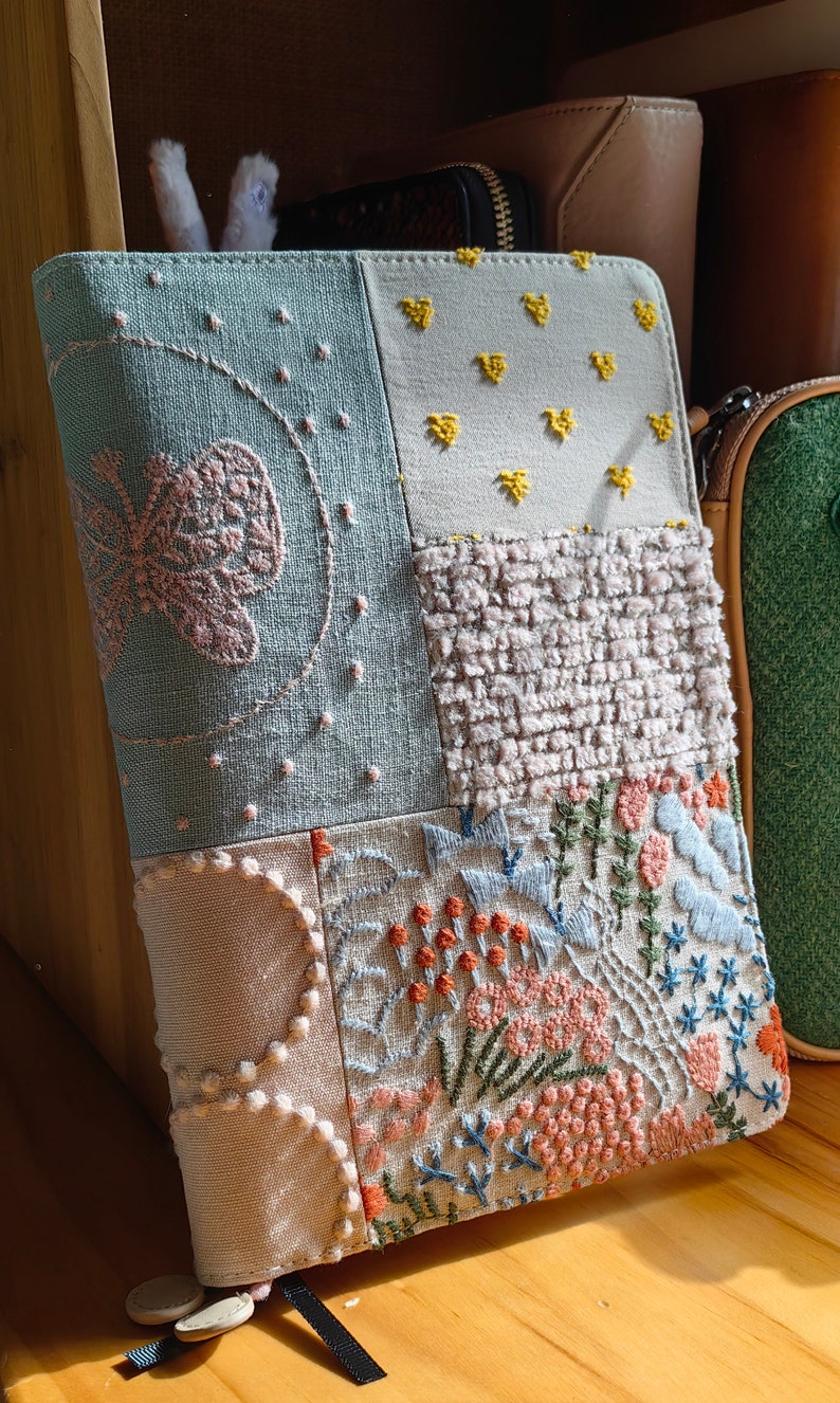 May include: A patchwork fabric notebook cover with embroidered designs. The cover features a butterfly, flowers, and geometric patterns in shades of blue, pink, yellow, and orange.