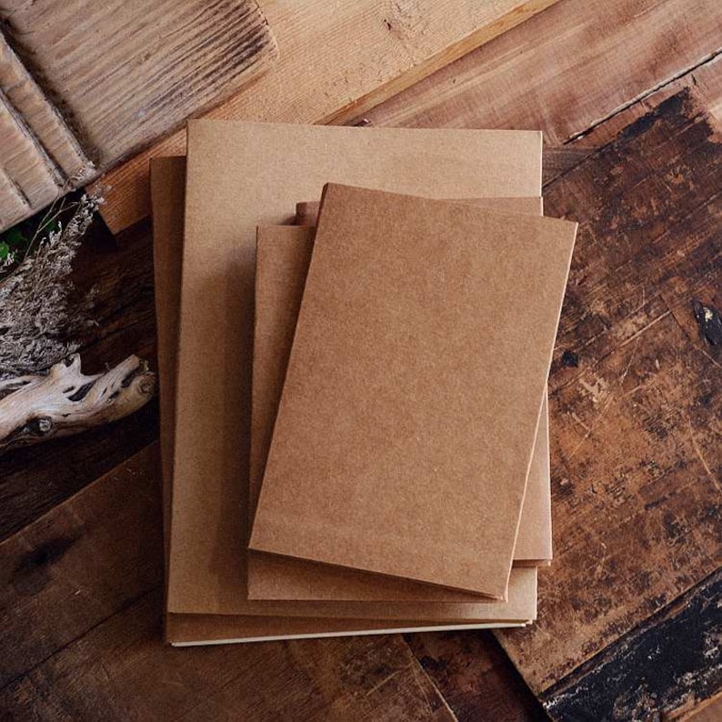 May include: A stack of four brown kraft paper notebooks with blank covers. The notebooks are arranged in a staggered pattern on a wooden surface.