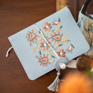 May include: A pale blue journal featuring an embroidered floral pattern in orange, teal, and gold. It has a tassel and pearl closure. The cover is made of textured fabric, and it's secured with a string.