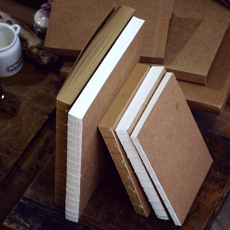 May include: Four brown notebooks with white covers stacked on top of each other. The notebooks are bound with a stitch and have a rough texture.