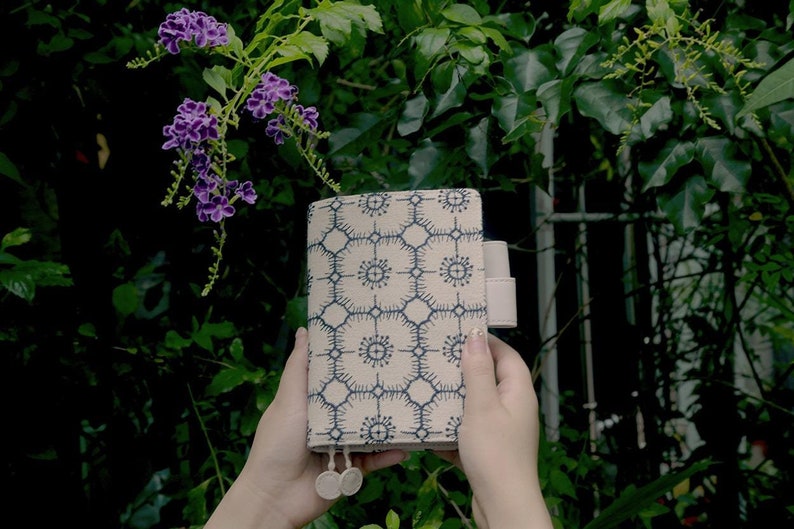 Op de afbeelding: Een beige stoffen notitieboekomslag met een geometrisch patroon in blauw en wit. De omslag heeft een klep sluiting en een band.