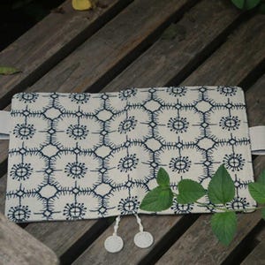 Op de afbeelding: Een witte stoffen notitieboekomslag met een blauw geometrisch patroon en witte kwastsluitingen. De omslag ligt op een houten oppervlak.