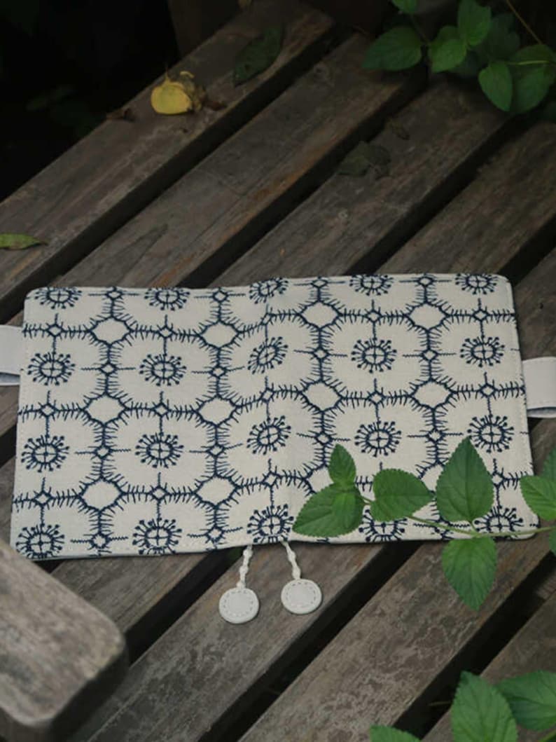 Op de afbeelding: Een witte stoffen notitieboekomslag met een blauw geometrisch patroon en witte kwastsluitingen. De omslag ligt op een houten oppervlak.