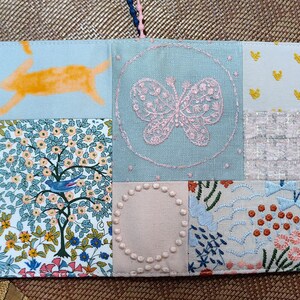 May include: A light blue fabric notebook cover with a patchwork design. The cover features a variety of embroidered designs, including a butterfly, a bird, and a rabbit. The cover is made of different fabrics, including a floral print, a solid blue fabric, and a white fabric with a small pattern.