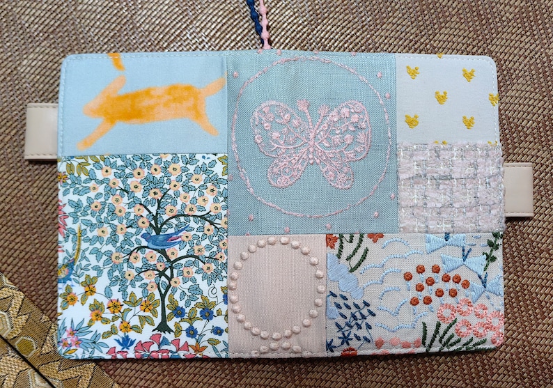 May include: A light blue fabric notebook cover with a patchwork design. The cover features a variety of embroidered designs, including a butterfly, a bird, and a rabbit. The cover is made of different fabrics, including a floral print, a solid blue fabric, and a white fabric with a small pattern.