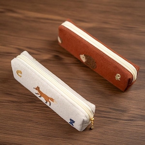 Op de afbeelding: Twee rechthoekige etuis met crèmekleurige ritsen. De ene etui is lichtbeige met een geborduurde vos en kleine blauwe en gouden accenten. De andere etui is roestkleurig met een geborduurde egel en kleine gouden accenten.