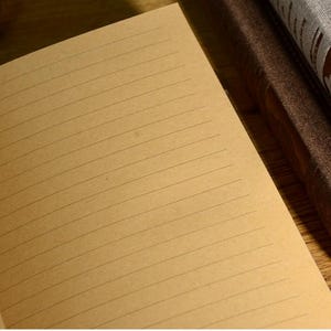 以下が含まれることがあります： 罫線入りのベージュ色のページが開いたノート。ページはわずかに傾いており、背景にある茶色の本の背表紙が垣間見える。線は均等に配置されており、クラシックなノートのデザインを示唆している。