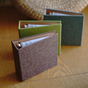 May include: Three fabric-covered photo albums in brown, green, and olive green. The albums are closed and have a clear plastic window on the front cover.