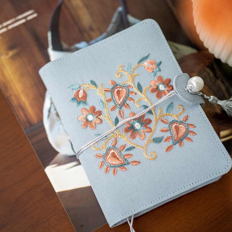May include: A pale blue journal featuring an embroidered floral pattern in orange, teal, and gold. It has a pearl clasp and a grey tassel. The cover is made of a textured fabric.