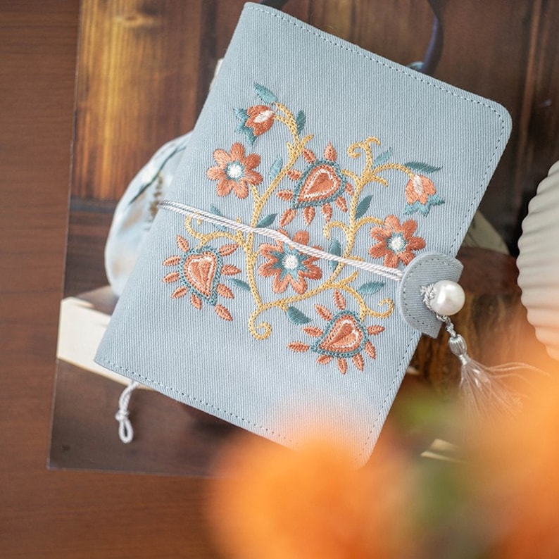 May include: A pale blue journal cover featuring an embroidered floral pattern in orange, peach, and gold. The cover is fastened with a white cord and a grey button with a pearl and tassel. The journal is on a wooden surface.