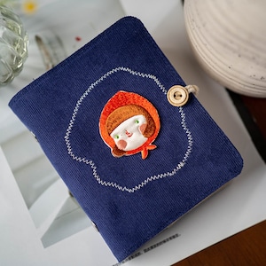 Peut inclure: Un petit carnet en velours côtelé bleu avec une bordure cousue et une jolie fille brodée avec une capuche rouge. Le carnet a une fermeture à bouton en bois.