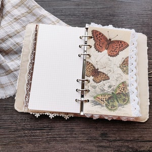 May include: A brown leather bound journal with white lace trim and a page featuring a vintage illustration of butterflies. The journal has a ring binder and a blank page.
